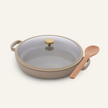 Beige Dual Handle Always Pan with nonstick finish and lid featuring a gold knob, accompanied by a wooden spoon, displayed on a neutral background.