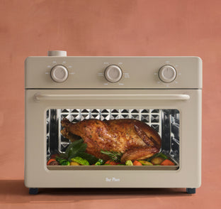 Beige toaster oven with a roasted chicken and vegetables inside on a peach background