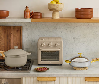A kitchen countertop with a beige air fryer, a beige pot on the stove, a lidded pan, a plate of grapes, and various ceramic dishes and bowls on wooden shelves.