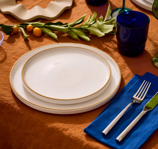 A table set with two white plates with gold rims, a fork and knife on a blue napkin, and decorative greenery on an orange tablecloth.