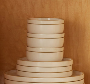 Stack of cream-colored ceramic bowls and plates with thin brown rims, arranged against a wooden background.