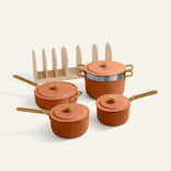 The Essentials Saucepan Set includes four orange pots and pans with lids, gold handles, and non-toxic nonstick surfaces. Shown with a beige rack, these pans nest together for convenient storage.