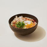A bowl of rice porridge topped with shredded chicken, a soft-cooked egg, sliced green onions, and seasoning.